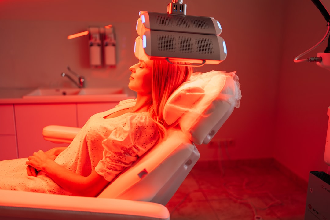 about-04 Side view portrait of blonde female lying on procedure chair having LED red light hair photodynamic therapy during hair treatment. Woman client having non-invasive type of phototherapy.